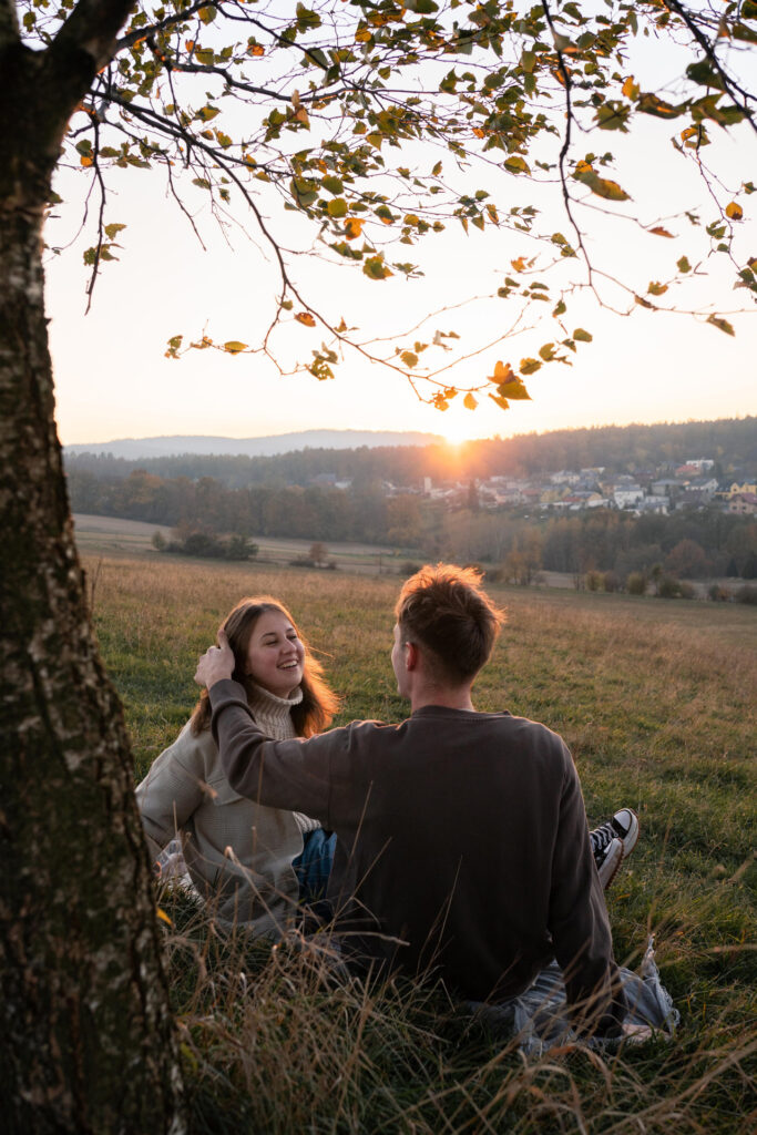Mladý pár sedí na louce u stromu při západu slunce, romantická atmosféra.