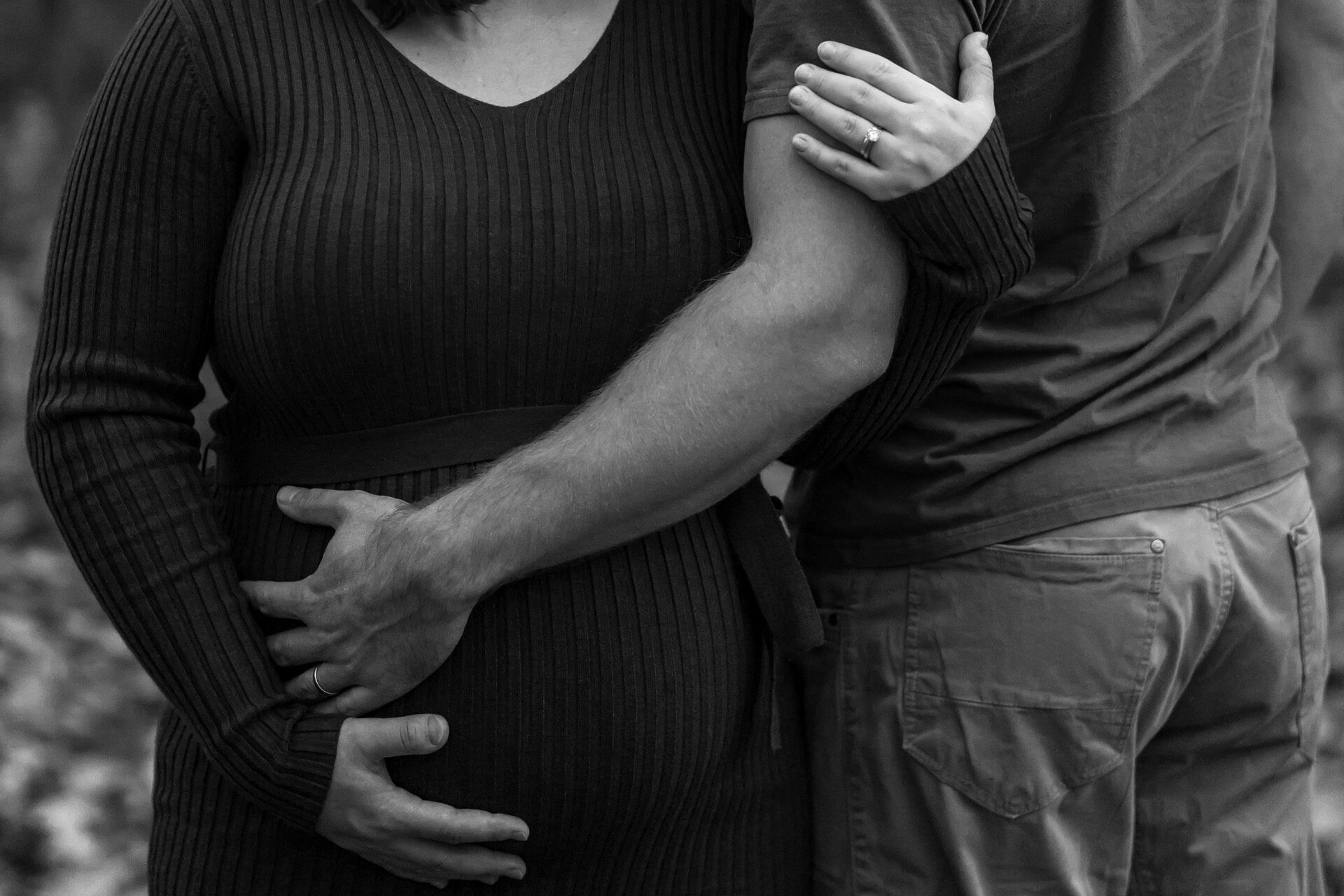 Detail těhotenského bříška v objetí partnera, černobílá fotografie.