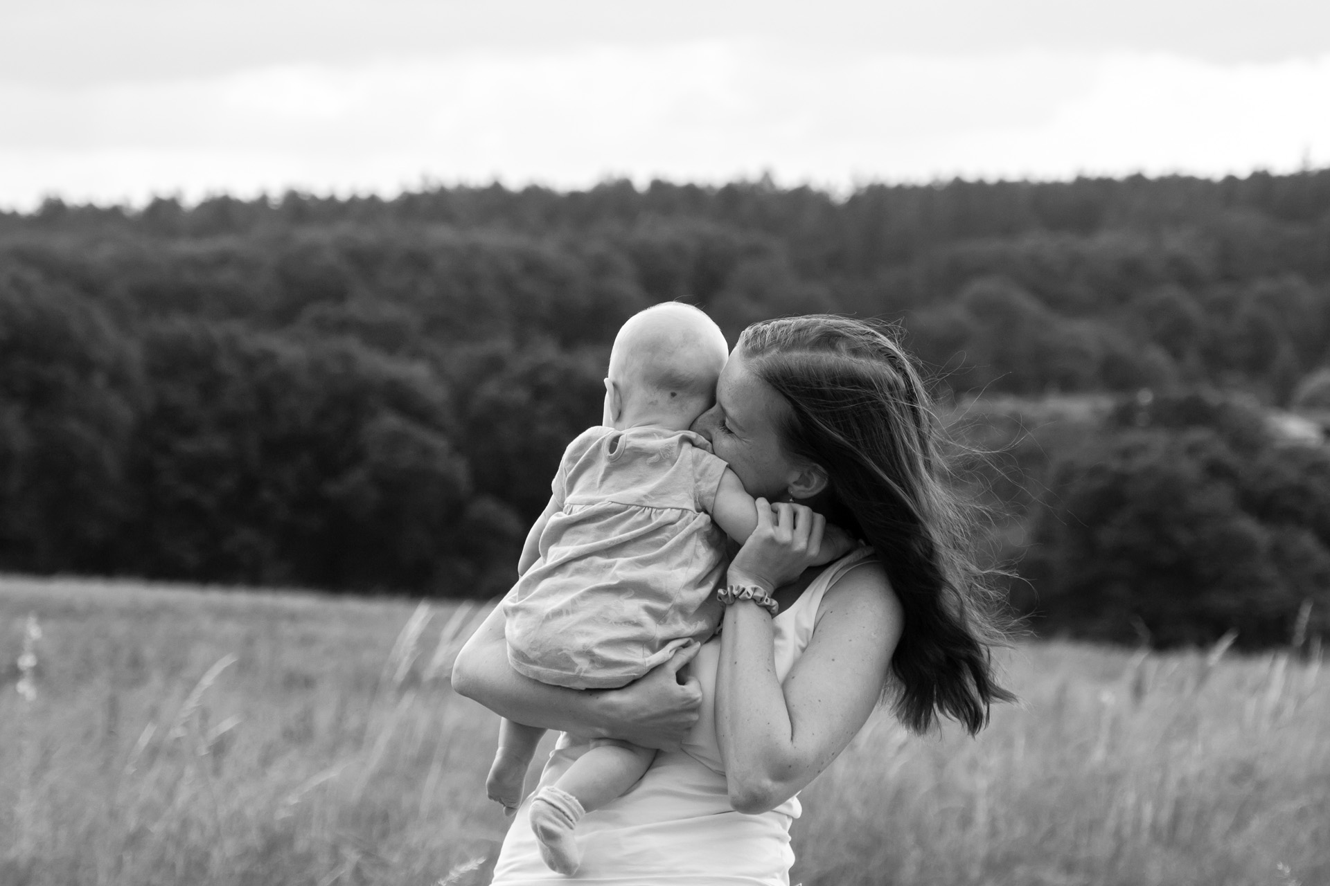 Žena nese dítě v náručí a kráčí po louce, vítr ve vlasech.