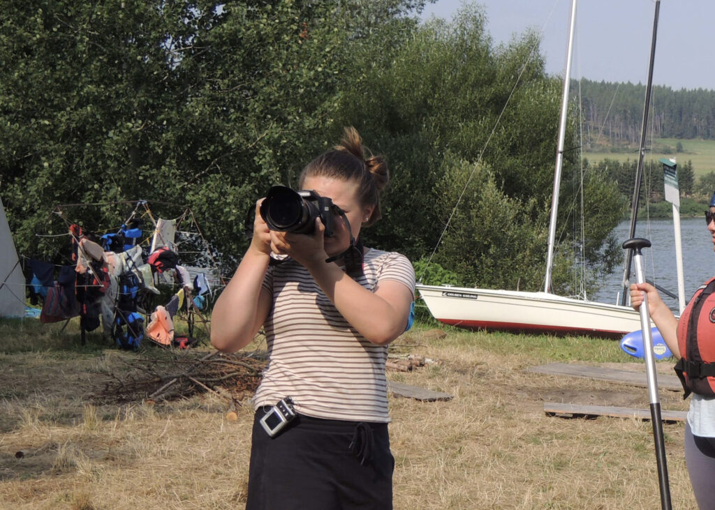 Mladá dívka drží fotoaparát u vody, zaměřuje objektiv.